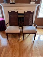 Pair of two traditional wooden armchairs with cane backs and beige upholstered seat cushions, shown together in front of a fireplace on wooden floor.