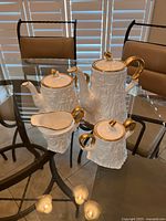 Full set of four pieces displayed on glass table, showing coffee pot, tea pot, creamer, and sugar bowl with gold accents and detailed bark texture.