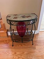 View of small accent table showing the tile-like top with leaf and vine pattern, metal frame and lower shelf holding decorative objects.