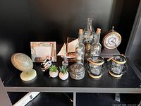 Photo showing a shelf with various small decorative items including a vintage clock, glass oil lamp, miniature sailboat, globe, plants, metal container, and lanterns