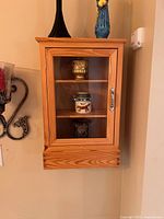 Wooden wall-mounted cabinet shown closed with decorative pots and vases visible through the glass door and on top of the cabinet.