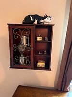 Front view of small dark wood wall-mounted cabinet with glass door featuring metal scrollwork, containing decorative metal pitchers and container, open shelves with small trinket box, bird figure, glass figurines, and colorful tin box, with black and white cat figurine on top.