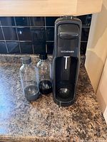 Front view of black SodaStream machine with two bottles on a kitchen counter, one bottle with gray cap and one bottle with black cap
