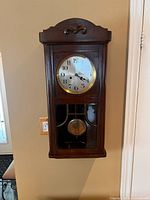 Full view of vintage wooden wall clock mounted on wall showing clock face and pendulum behind glass.