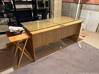 Wood desk shown from side angle with separate glass top piece loosely resting on it; side table next to desk, carpeted floor, basement setting