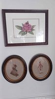 Overall view of three framed wall art pieces hung on a wall showing two oval needlepoint art and one rectangular floral print.