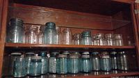 Shelf displaying a collection of approximately 20 blue and clear antique glass jars of various sizes, some with lids and some without.