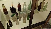 Large assortment of antique and vintage bottles in various glass colors including clear, brown, green, and amber, shown inside a storage cabinet.