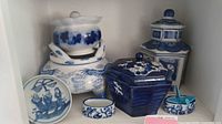 Seven pieces of blue and white oriental porcelain ceramics arranged on a shelf; includes lidded jars, dishes, and containers with blue painted Oriental designs.