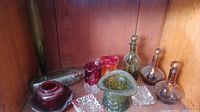 Various vintage and antique glass pieces including decanters, vases, and ashtrays in amber, green, clear, and ruby colors displayed on wooden surface.