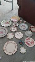 Photo showing multiple vintage decorative plates and cups arranged on carpet. Various sizes and patterns, mix of floral hand-painted designs and gold trim.