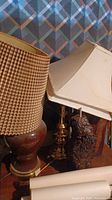 Photo showing three table lamps with various wooden and brass bases and plain lampshades, one fabric lampshade damaged.