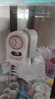 White ceramic clock showing Roman numerals and floral painted details with glass pendulum window.