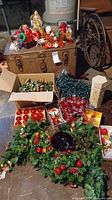 Wide image showing boxes with assorted Christmas ornaments, greenery garland with red berries, boxes of Christmas lights, and a brown trunk storing additional decorations.