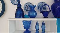 Seven pieces of cobalt blue vintage pressed glassware on a white shelf including a bell, pitcher, compote, basket, salt & pepper shakers, and a ruffled dish.