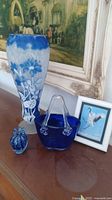 Wide view showing tall blue and white etched glass vase, small blue basket vase, blue glass paperweight, and framed mini art with bird.