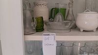 Shelf with various glassware and ceramic pieces including green glass pitcher, clear glass pitcher, juicer, decanter, and white footed bowls.