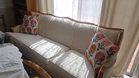 Full view of white upholstered sofa seating three, with wood frame and floral patterned throw pillows, sunlight casting shadows through curtains.