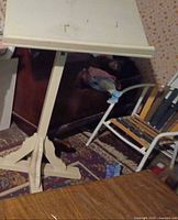 White wooden adjustable podium stand with an angled top surface showing slight wear and two metal hooks, supported by a four-legged base with crossbars, pictured in a room with patterned carpet and various chairs.