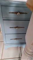 Front view showing three drawers with metal drawer pulls with white ceramic centers, painted light blue wood with minor wear.