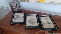 Wide view of 3 framed nautical ink drawings and octagonal tin sitting on wooden surface