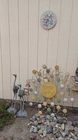 Two metal stork statues standing on ground next to a yellow metal sun sculpture surrounded by small garden rocks.