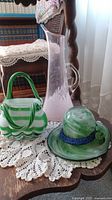 Photo showing the pink blown glass pitcher along with green/white decorative basket and hat placed on a crochet doily on a wooden table.