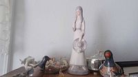 Porcelain lady figurine standing upright, white/grayish dress, holding a large round hat, surrounded by other figurines.