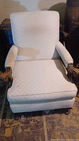 Front view of upholstered armchair showing patterned light cream fabric and carved wood arms with line of wooden legs beneath.