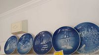 Five Royal Copenhagen blue and white porcelain Christmas collector plates displayed in a row, showing their detailed winter scenes and round shape.