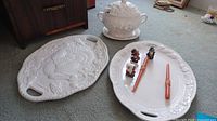 White ceramic soup tureen with lid and two large white ceramic serving platters on carpeted floor, one white platter embossed with turkey and fruit design, other platter plain white with fruit components.