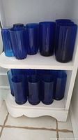Set of 12 cobalt blue faceted glass drinking glasses arranged on white shelves in various positions, showing the color and shape variations