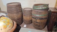 Three wooden barrels of varying sizes arranged together on the floor near a globe and furniture. Visible aged wood and metal bands, some green paint on one barrel's bands.