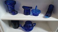 Set of six cobalt blue glass miniature collectibles shown on white shelves, featuring boots, a top hat, basket, cup, and teapot shapes.