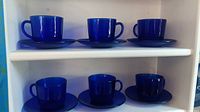 Six blue glass teacups arranged on two shelves with matching saucers beneath each cup.