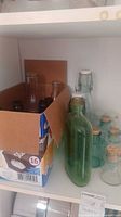 Shelf containing various clear, green, and dark brown glass bottles with corks and swing-top stoppers in a cardboard box and next to it.