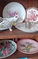 Photo of vintage ceramic and porcelain plates with floral and grape motifs, a porcelain basket, and a small metal miniature pitcher.