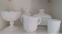 Front view of four milk glass items grouped: footed bowl, lidded covered dish, lidded cup, and tumbler.