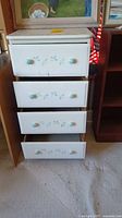 Front view of the white wooden chest showing four drawers with blue/green floral detailing and round knobs. Bottom drawer is open.
