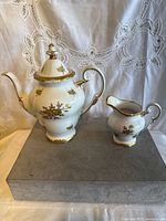Photo of white porcelain teapot and matching creamer with gold floral design and gold trim shown on grey box against lace curtain background