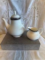 White teapot with curved spout and lid with knob, along with matching milk bowl. Both pieces decorated with black and silver band near rim.