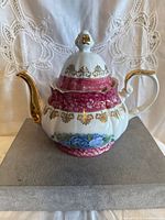Front left angle view of vintage porcelain teapot showing pink floral band, gold gilding on handle and spout, and blue flower decoration on body.