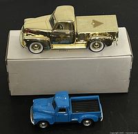 Side and above view of gold 1949 Chevy pickup metal bank with '2000' and Canadian Tire logo on door, placed on box, next to a small blue die cast pickup truck.