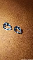 Two pear shaped light blue aquamarine gemstones displayed on a brown textured surface.