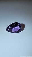 Pear cut purple amethyst gemstone photographed under lighting showing facets and deep purple color on white background.