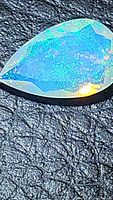 Close-up of pear-shaped faceted natural opal showing bright blue and green pinfire color pattern with yellow reflections on black textured surface.