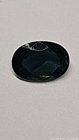 Close-up photo of a natural green sapphire, oval cut, showing dark green transparent faceted surface and slight inclusions.