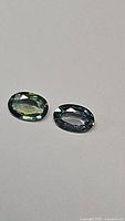 Pair of natural green sapphires oval cut lying flat on a white surface showing facets and clarity in detail.