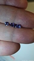 Four deep blue cushion cut tanzanite gemstones placed on a fingertip showing size relative to a finger. The stones are VVS-flawless, approximately 3.8mm each.