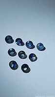 Photo of ten natural multicolored sapphire gemstones arranged on white surface showing various blue and green hues.